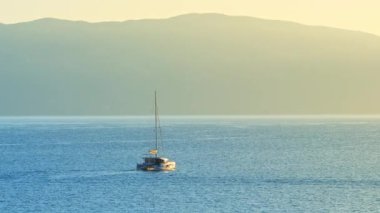 Gündoğumunda Yunanistan, İyon Denizi 'nde yelkenli. Dağın arka planına karşı denizde yüzen bir tekne. Yelkenli teknesi katamaran, Kefalonya adası, Yunanistan. Hala vurulmuş. 