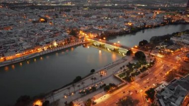 Aerial view of the Seville cityscape at night, Andalusia region, Spain. Flying over Guadalquivir river with historic Triana Bridge in Seville old town with night city illumination. 