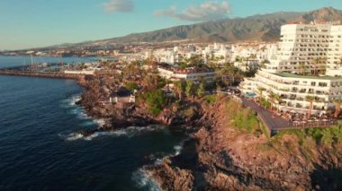 Costa Adeje tatil beldesi üzerinde gün batımında Tenerife, Kanarya Adaları, İspanya. Lüks otelleri, palmiye ağaçları ve arka planda güzel dağları olan Tenerife adasının kayalık sahili.