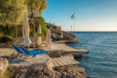 Kayalıklarda şemsiyeli güzel küçük plaj ve Yunanistan 'ın İyon Denizi' ndeki Kefalonia Adası 'nda iki boş güneşlik. Mükemmel bir yaz tatili yeri. Arka planda Yunan ulusal bayrağı