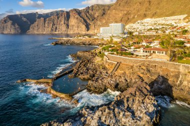 Günbatımında Tenerife adasındaki Los Gigantes manzarası, İspanya. Los Gigantes 'in doğal havuzu, lüks oteller, sahil kenti mimarisi ve arka planda görkemli yüksek uçurumlar, Tenerife, İspanya