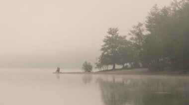 Sisli bir sabahta göl kıyısında balık tutan insanların silueti. Sisli gölün yoğun sonbahar sisinin içinde kaybolduğu sakin, minimalist bir manzara. Sakin ve zen benzeri bir sahne.