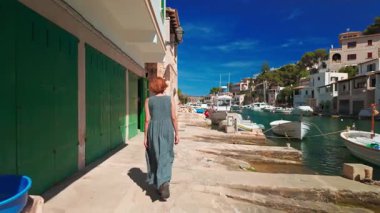 Uzun yaz elbiseli bir kadın, İspanya 'nın Balearic Adaları yakınlarındaki Santanyi, Mallorca yakınlarındaki Cala Figuera adlı büyüleyici balıkçı köyünde yürüyor. Geleneksel Akdeniz tarzı kıyı evleri yakınlarında demirlemiş küçük tekneler.