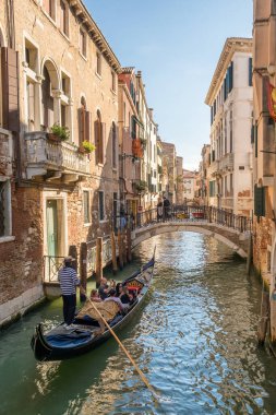 Venedik, İtalya - 11 Ekim 2024: Venedikli gondolcu Venedik, İtalya 'da dar bir kanal boyunca turistlerle gondolda yolculuk ediyor.