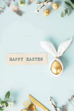 Stylish Easter flat lay with golden egg in easter bunny napkin, golden quail eggs, feathers, cutlery and spring flowers on blue background. Minimalist modern Easter table decorations. Top view flat
