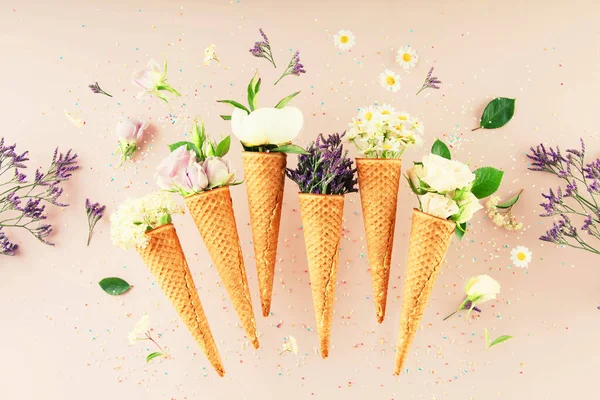 Flat-lay of waffle cones with flowers over pastel light pink background, top view. Spring or summer mood concept
