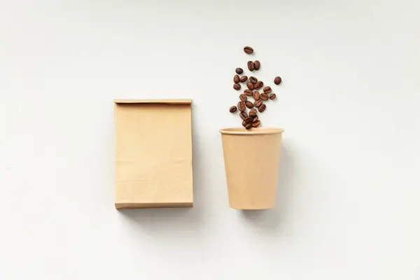 A paper coffee cup beside a brown bag with coffee beans on a white background.