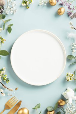 Easter table decorations. Happy Easter concept with white plate, golden easter eggs, feathers and spring flowers on blue background. Flat lay top view copy space