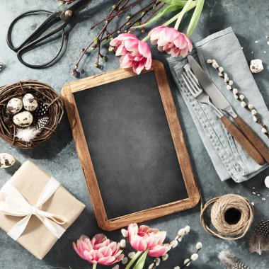 Easter table setting with vintage chalk board, spring flowers and cutlery on dark blue background top view flat lay mock up