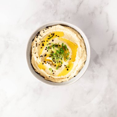 Üzerinde siyah susam tohumları ve taze mikroyeşillikler bulunan zeytinyağı serpilmiş bir kase pürüzsüz humus mermer zemin üzerinde servis edilir. Şık ve sağlıklı bir bitki tabanlı yemek..