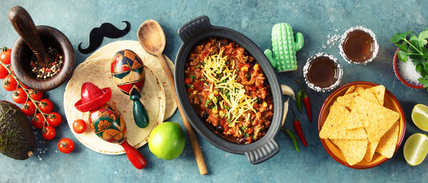 Top view of Mexican beef chili con carne topped with cheese, surrounded by nachos, tequila, maracas, and fresh ingredients on a vibrant blue background. Banner, header
