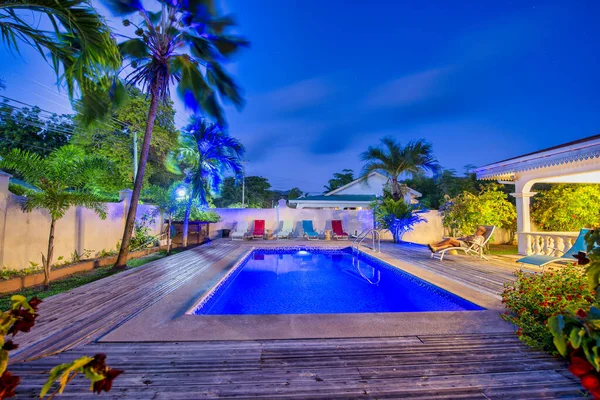 Night view of a tropical house with pool and palms.