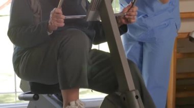 Yavaş çekimde Asyalı antrenör egzersiz bisikletinde bacak egzersizi yapan kadına yardım ediyor. Rehabilitasyon ve emeklilik kavramı.