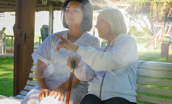 亚洲医生用手杖与年老的退休妇女交谈 她们都坐在花园的长椅上 幸福与退休概念 — 图库照片