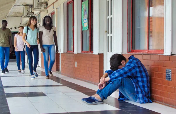 Young caucasian sitting alone with sad feeling at school. Child in depression abandoned in a corridor and leaning against brick wall. Bullying, discrimination and racism concept.
