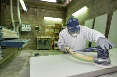 Bir erkek işçi maske takıyor ve verniklemek için ahşabı zımparalıyor..