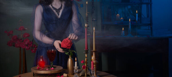 Young beautiful woman in blue vintage dress with red roses makind potion in dark room