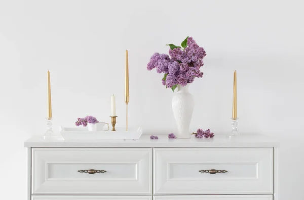 在白色花瓶中的丁香花和背景白墙上的金色蜡烛 — 图库照片