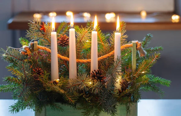 在家里的白色桌子上用冷杉树枝和四个燃烧的蜡烛装饰木制篮子 — 图库照片