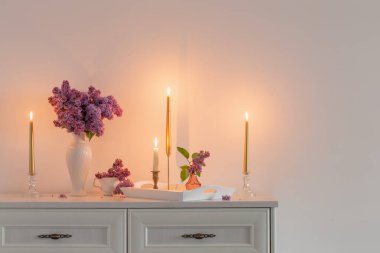 Beyaz vazodaki leylak çiçekleri ve arka plandaki altın mumlar. Beyaz duvar.