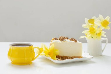 Bir fincan çay ve beyaz masada sarı bahar çiçekli cheesecake.