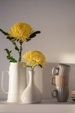 Vazoda sarı kasımpatı ve beyaz arka planda beyaz rafta duran fincanlar.