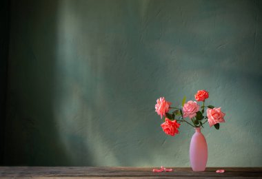 Pembe güller cam vazoda, ahşap masada, arka planda, karanlık duvarda.