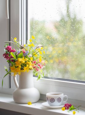 Beyaz sürahide yaz çiçekleri ve pencere kenarında bir fincan kahve.