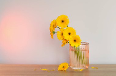 Sarı gerbera cam vazoda, ahşap masada, arka plandaki duvarda.