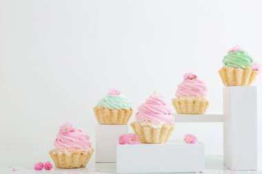 Beyaz arka planda tahta kürsülerde bahar çiçekleri olan pembe ve yeşil kapkekler.