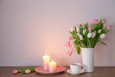 Bir fincan kahve ve vazoda bahar laleleri ve ahşap rafta yanan mumlar.