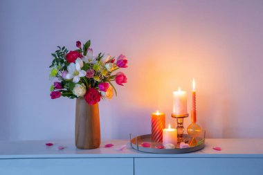 Vazodaki çiçekler ve arkadaki duvardaki rafta yanan mumlar.