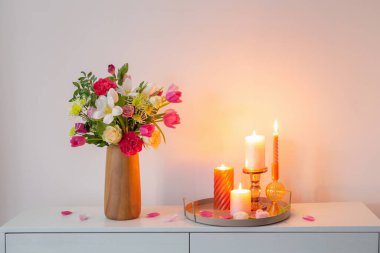 Vazodaki çiçekler ve arkadaki duvardaki rafta yanan mumlar.