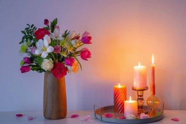 Vazodaki çiçekler ve arkadaki duvardaki rafta yanan mumlar.