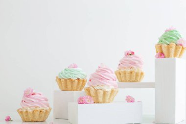 Beyaz arka planda tahta kürsülerde bahar çiçekleri olan pembe ve yeşil kapkekler.