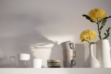 Vazoda sarı kasımpatı ve beyaz arka planda beyaz rafta duran fincanlar.