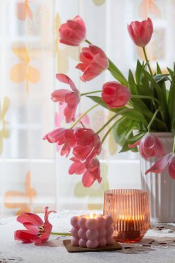Beyaz vazoda pembe laleler ve arka plan penceresinde yanan mumlar.