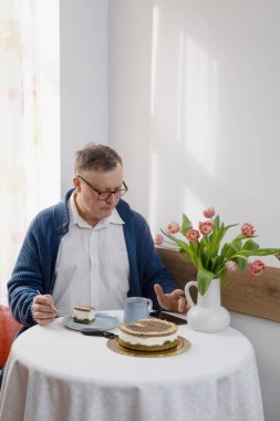 Yaşlı bir adam mutfağında akıllı telefonuyla pasta yiyor.