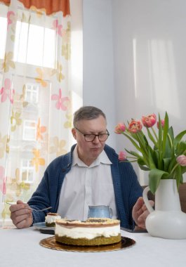 Yaşlı bir adam mutfağında akıllı telefonuyla pasta yiyor.