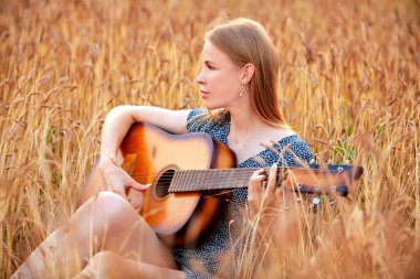 Güzel bir genç kadın buğday tarlasında oturup akustik gitar çalıyor..