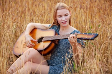 Güzel bir genç kadın buğday tarlasında oturup akustik gitar çalıyor.