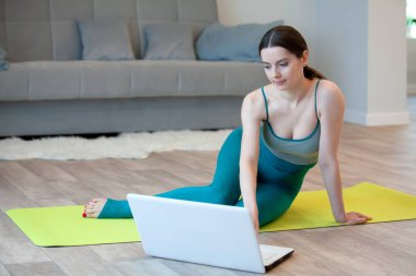 Spor kıyafetli genç bir kadın evde dizüstü bilgisayarla yoga yapıyor.