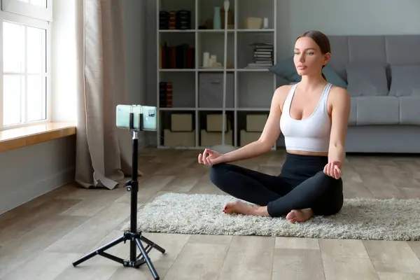 Kadın Evde Halıda Yoga Yapıyor Telefon Tripodla Lotus Pozisyonunda Oturuyor Telifsiz Stok Imajlar