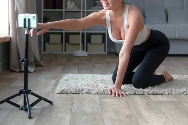 woman sitting on the floor in yoga pants recording an online fitness video with her phone and tripod, smiling at the camera in a living room setting