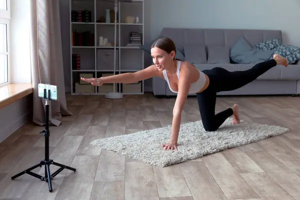 身穿黑腿和白色上衣的女性在家里的地毯上做瑜伽 用智能手机站在三脚架上为观众录制在线健身视频 免版税图库图片