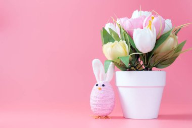 Pastel renkli Paskalya tavşanı süslemesi ve pembe arka planda dizilmiş bir saksı çiçek.