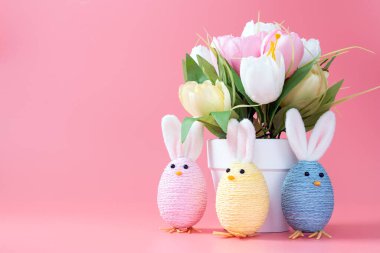 Pastel renkli Paskalya tavşanı süslemeleri ve pembe arka planda dizilmiş bir saksı çiçek.