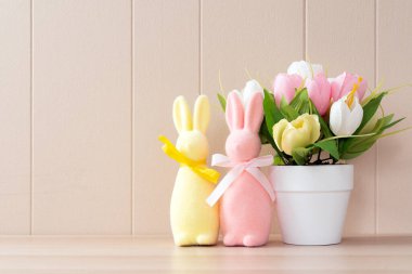 Pastel renkli Paskalya tavşanı süslemeleri ve beyaz arka planda dizilmiş bir saksı çiçek.