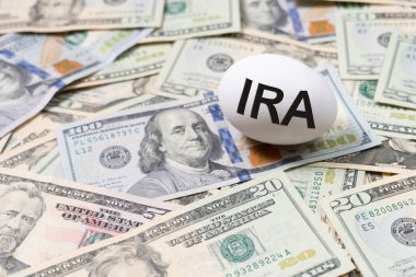 Bireysel Emeklilik Hesabı başlıklı bir beyaz yumurtayla emeklilik planlaması, IRA, yuva içi para metaforuyla temsil edilen ABD Doları banknotlarından oluşan bir yatağa yerleştirildi..