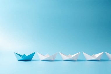 Mavi kâğıt gemiyle liderlik kavramı mavi arka planda izole edilmiş beyaz tekneler arasında öncülük ediyor.